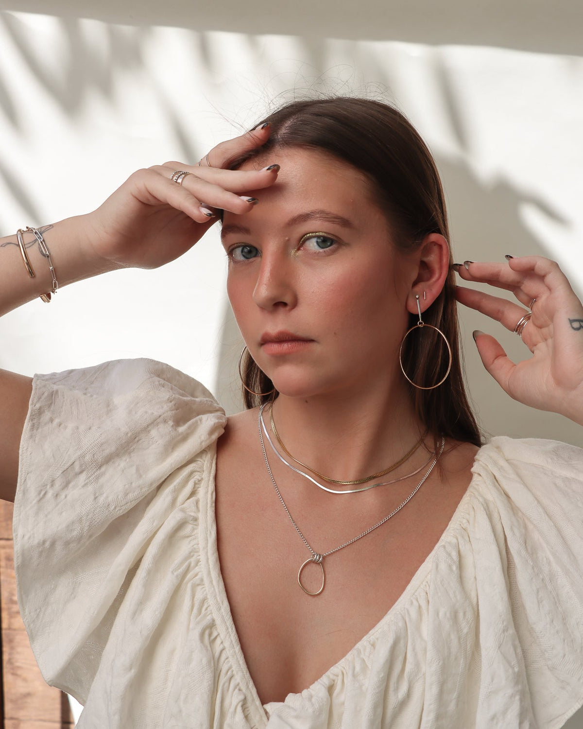 Model wearing mixed-metal layered necklaces, gold hoop earrings, delicate rings, and stacked bracelets with a white puff-sleeve blouse against a softly sunlit background.