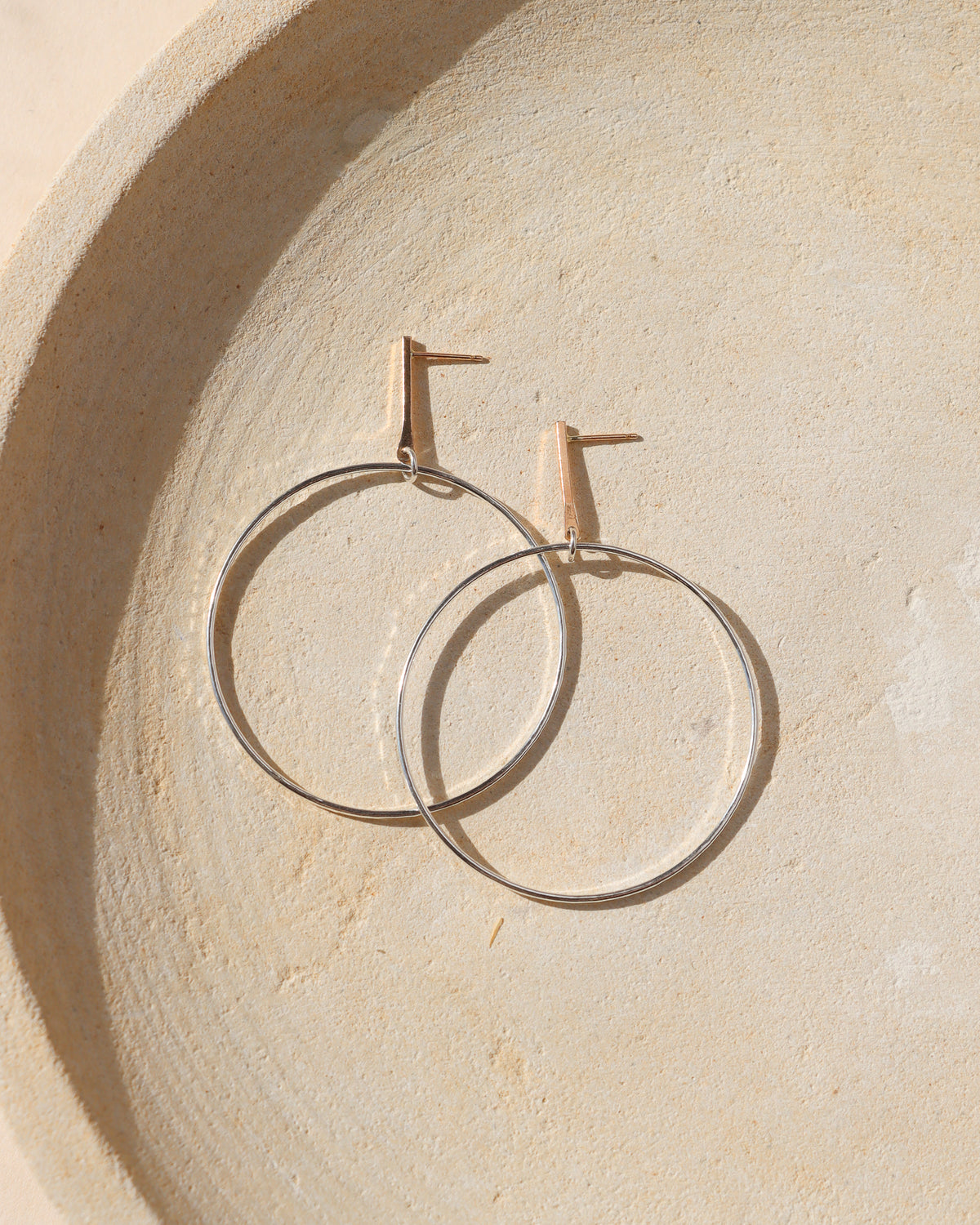 Silver hoop drop earrings with gold bar studs resting on a textured beige surface in soft sunlight.