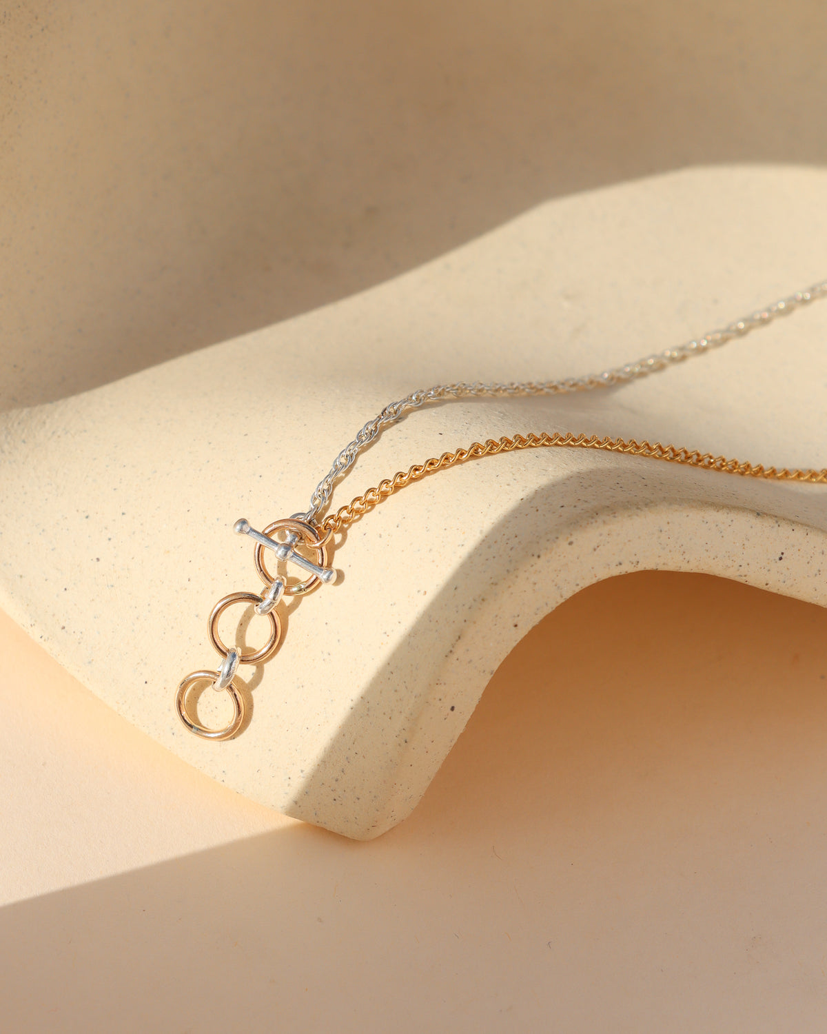 Alloy Toggle Necklace featuring gold fill and sterling silver chains joined by a front toggle clasp with dangling mixed metal rings, displayed on a cream ceramic surface in sunlight.