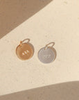 Two round angel number charms in gold and silver, stamped with 444 and 222, resting side by side on a textured cream surface in warm sunlight with soft shadows.