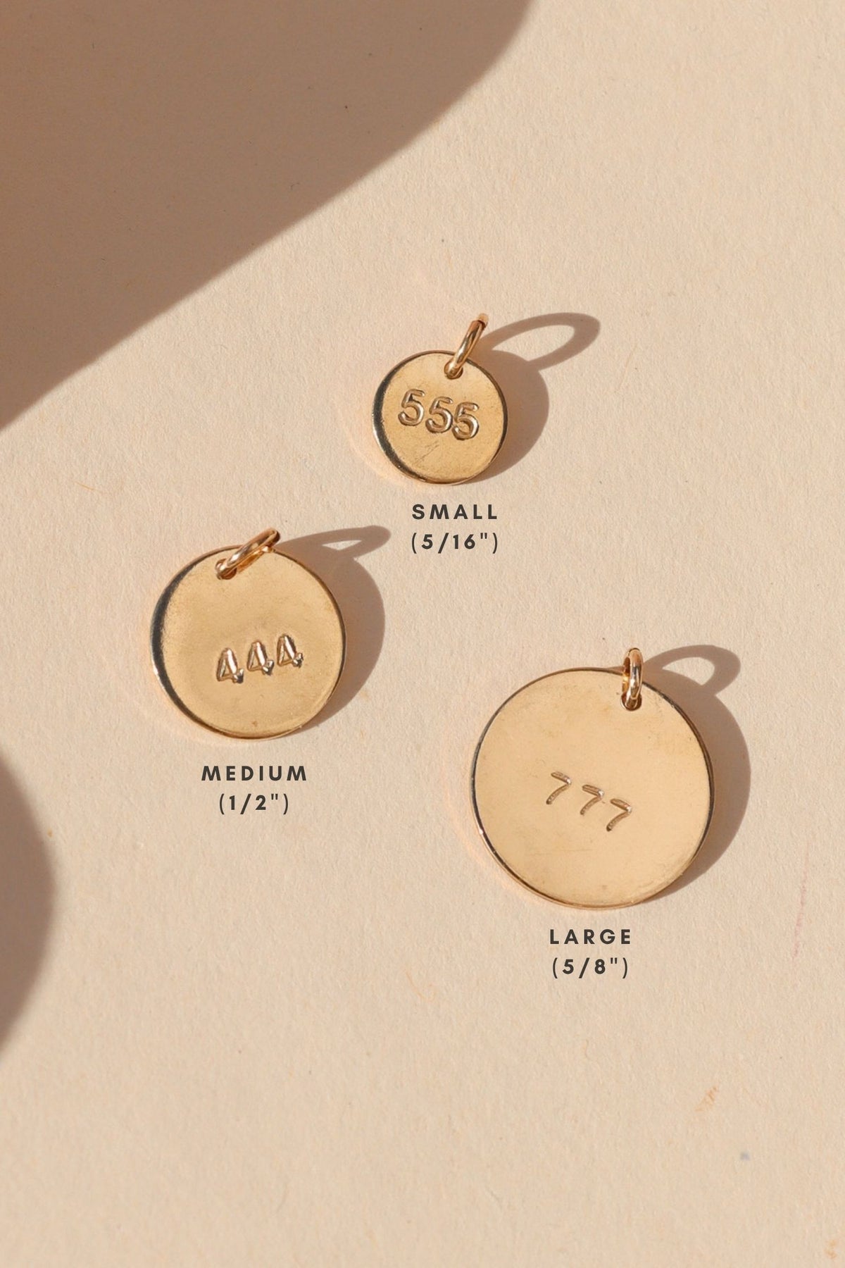 Three round gold number charms stamped with 555, 444, and 777, shown in small (5/16"), medium (1/2"), and large (5/8") sizes on a warm cream background with soft sunlight and shadows.