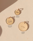Three round gold number charms stamped with 555, 444, and 777, shown in small (5/16"), medium (1/2"), and large (5/8") sizes on a warm cream background with soft sunlight and shadows.
