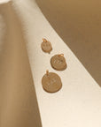 Three round gold angel number charms stamped with 111, 777, and 333 in graduated sizes, arranged on a textured cream surface with warm sunlight and soft shadows.