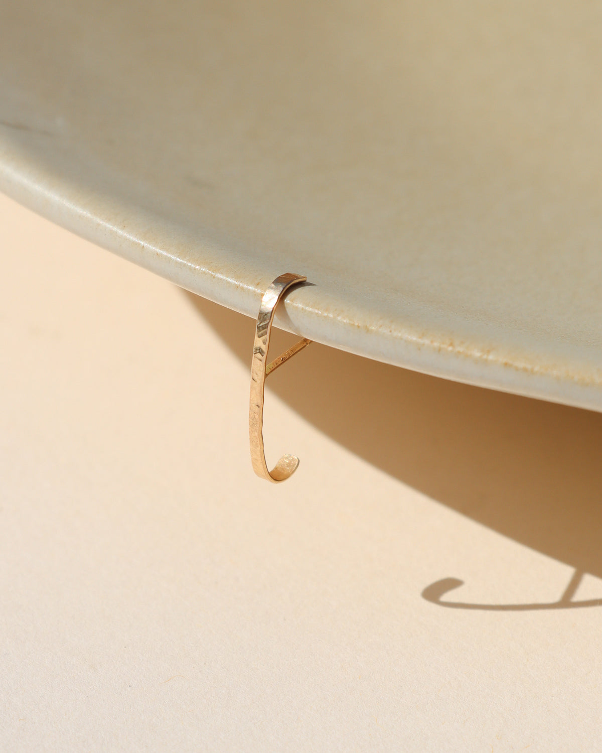 A delicate, thin gold huggie hoop earring draped over the edge of a light beige ceramic plate on a matching beige surface, casting a soft shadow.
