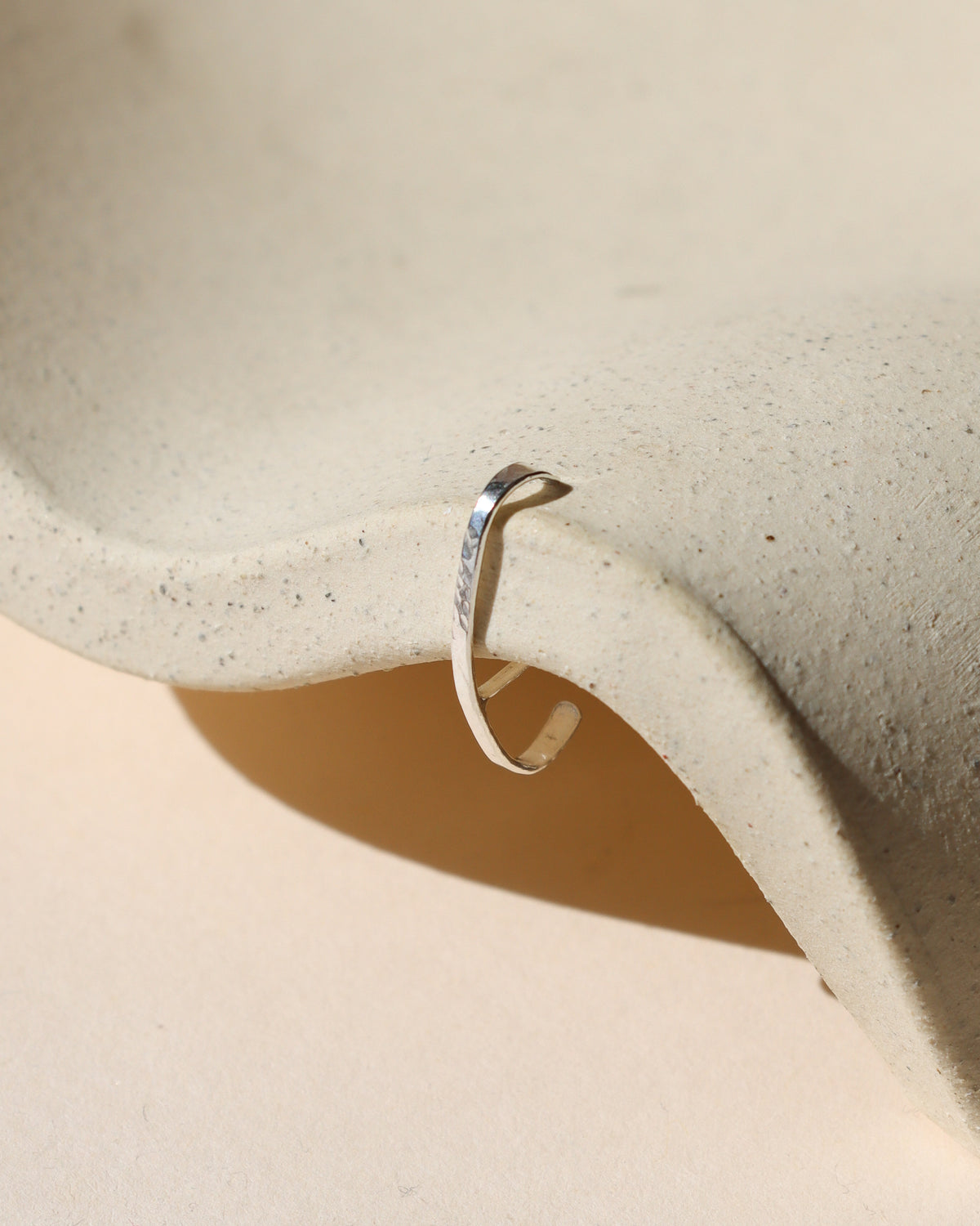 Hammered silver cuff earring with a stud detail, displayed on a textured cream ceramic form in warm sunlight.