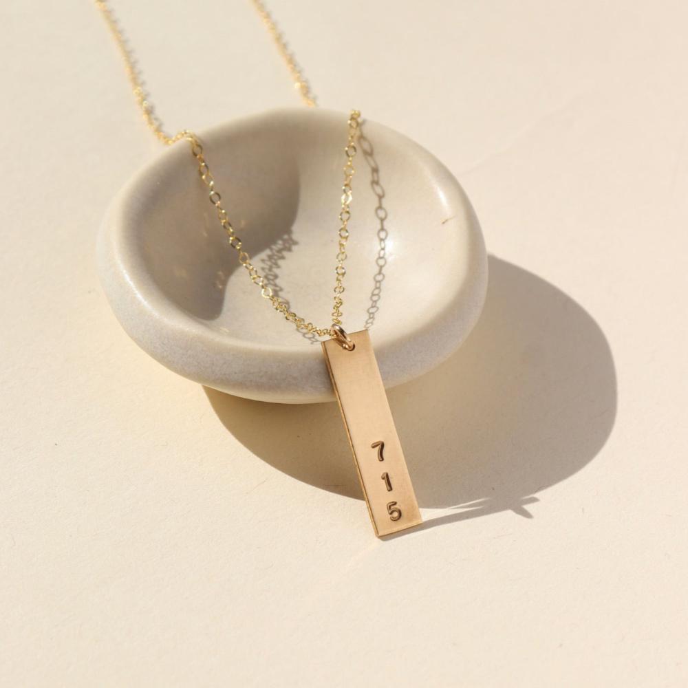 A gold hand-stamped, area code necklace with a delicate chain. Displayed on a small dish against a cream backdrop. 