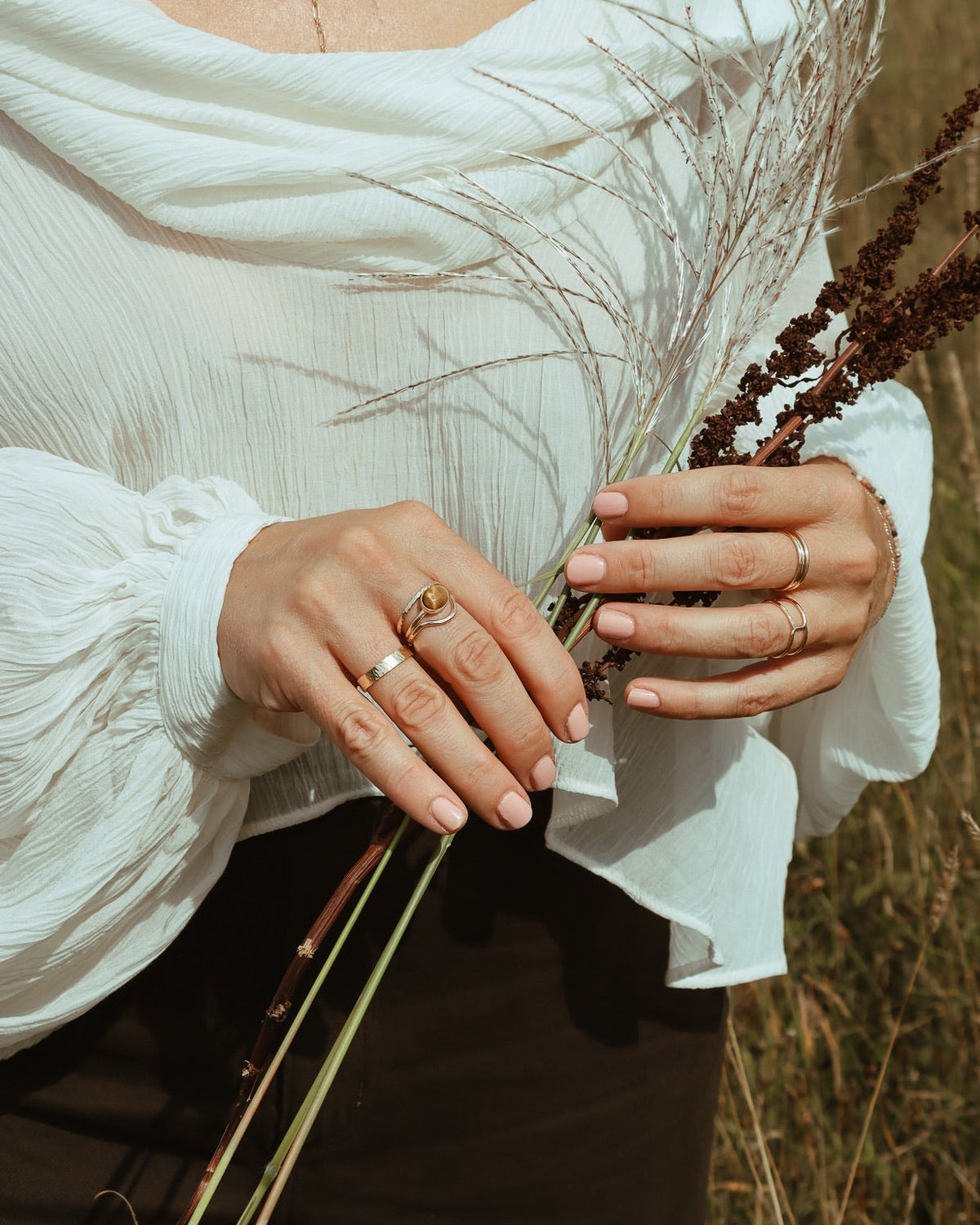 Hands wearing layered gold filled rings, including a textured band and oval gemstone ring, while holding dried grasses.
