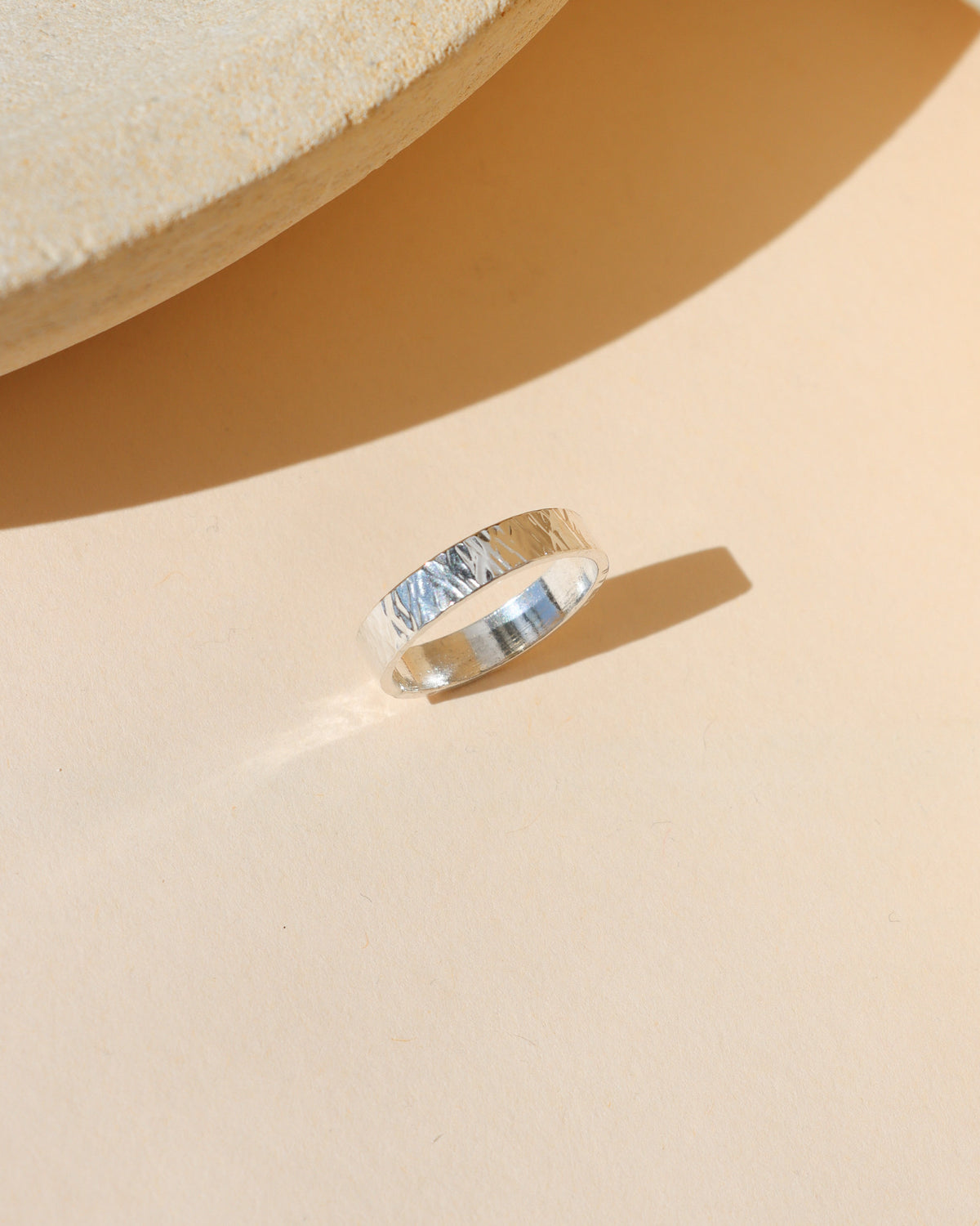 Wide silver band ring with a hand-textured finish, displayed in warm sunlight on a neutral background.