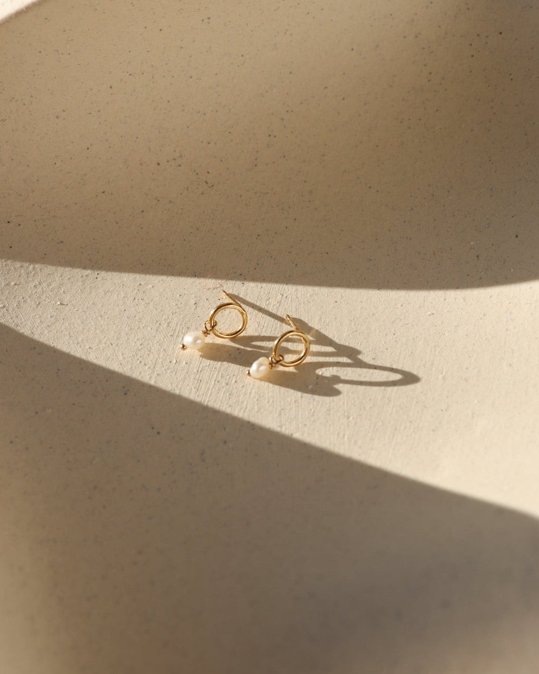 Product photo of 14k gold fill circle stud earrings featuring freshwater pearl drop accents, displayed on a textured ceramic surface in warm natural sunlight.