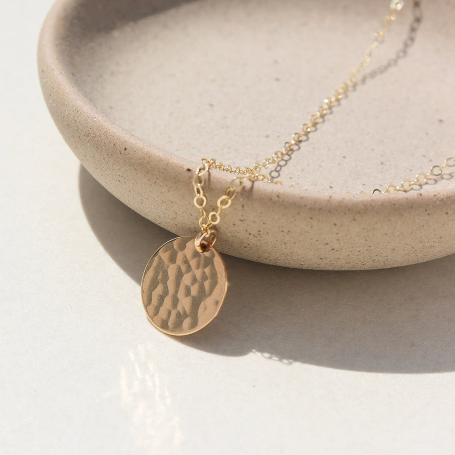 14k gold fill Moon drop necklace laid on a plate in the sunlight. This necklace features our simple chain met by a hammered coin, making it look like the moon.