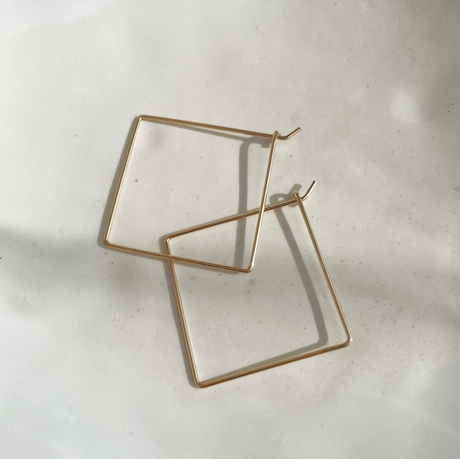 14k gold fill Square Hoops laid in the sunlight on a gray jewelry plate. These earrings feature a lightweight statement piece. - Token Jewelry