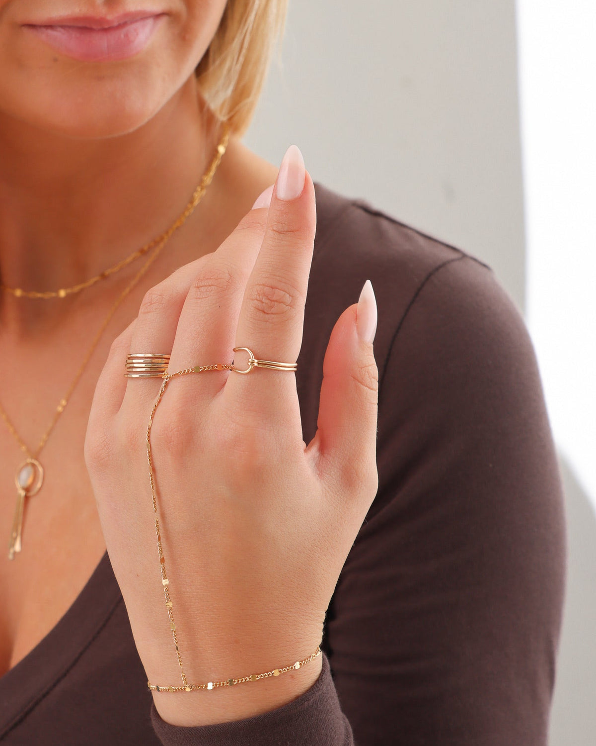 Close-up of a woman wearing a delicate gold Barrel Ring stacked with other gold rings, a draped gold hand chain bracelet, and layered gold necklaces with an opal pendant.
