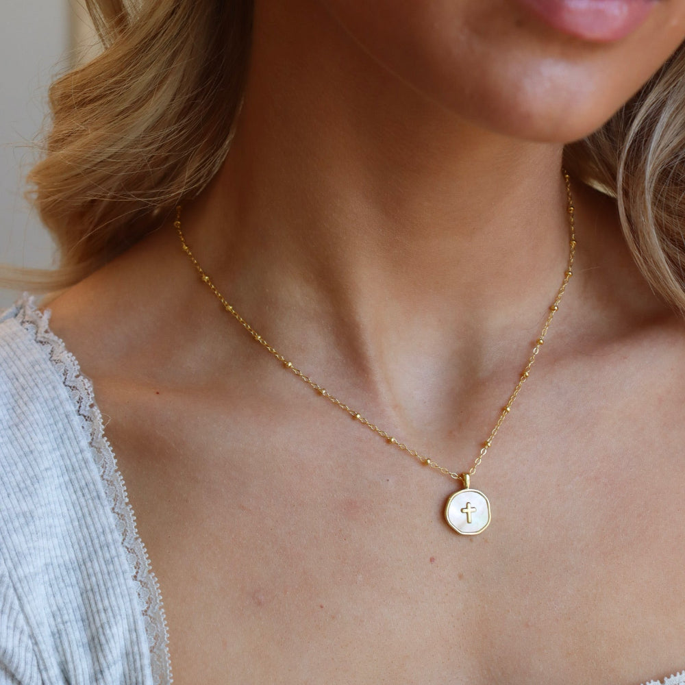 Model wearing a small beaded 14k gold fill chain featuring a circle mother of pearl cross charm pendant. 