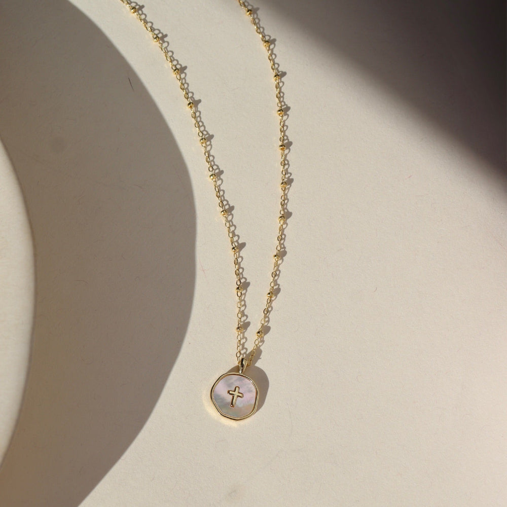 A small beaded 14k gold fill chain featuring a circle mother of pearl cross charm pendant. The necklace is displayed on a cream backdrop. 