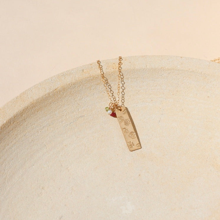 14k gold fill delicate chain necklace with hand stamped flowers on a bar pendant with birthstone charms on a light dish