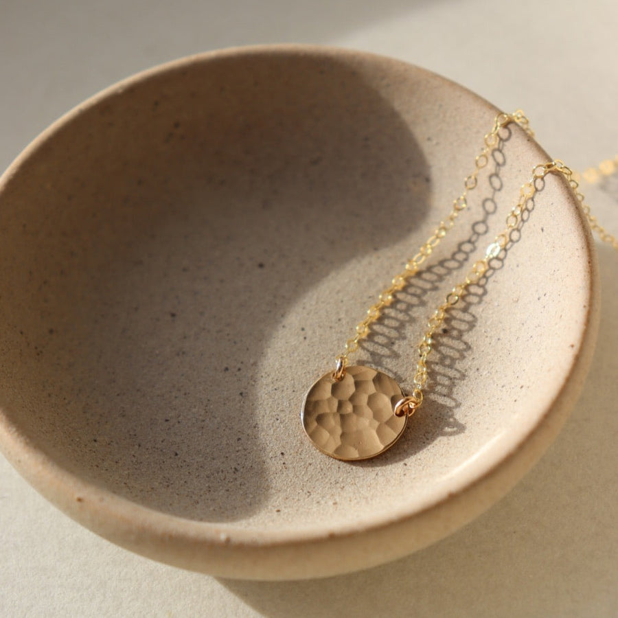14k gold fill Mini Moon Necklace laid in the sunlight on a gray plate. This Necklace features a disc that is completely hammered connected by a simple chain.