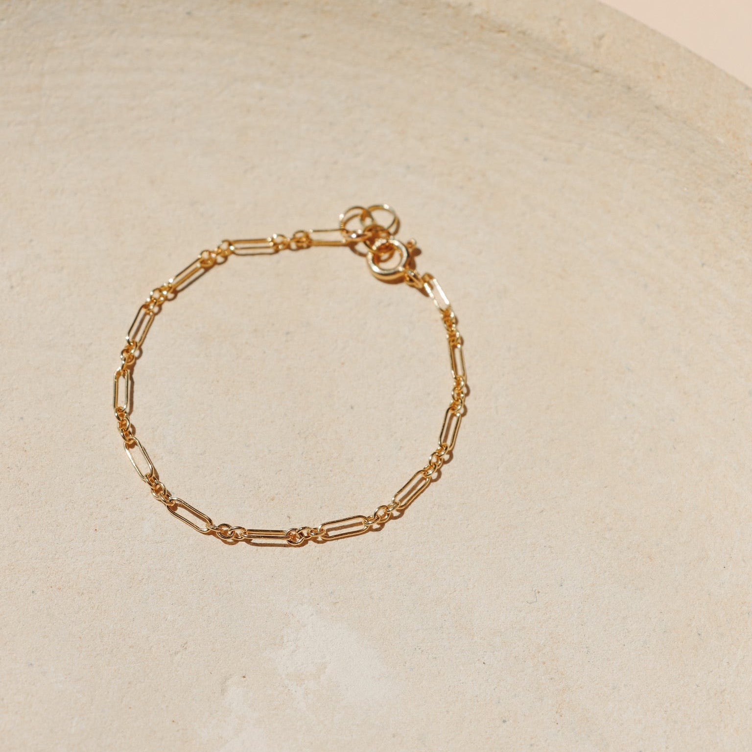 Minimalist gold chain bracelet with delicate elongated rectangular links and a spring ring clasp, arranged on a neutral, textured stone tray under soft natural lighting.