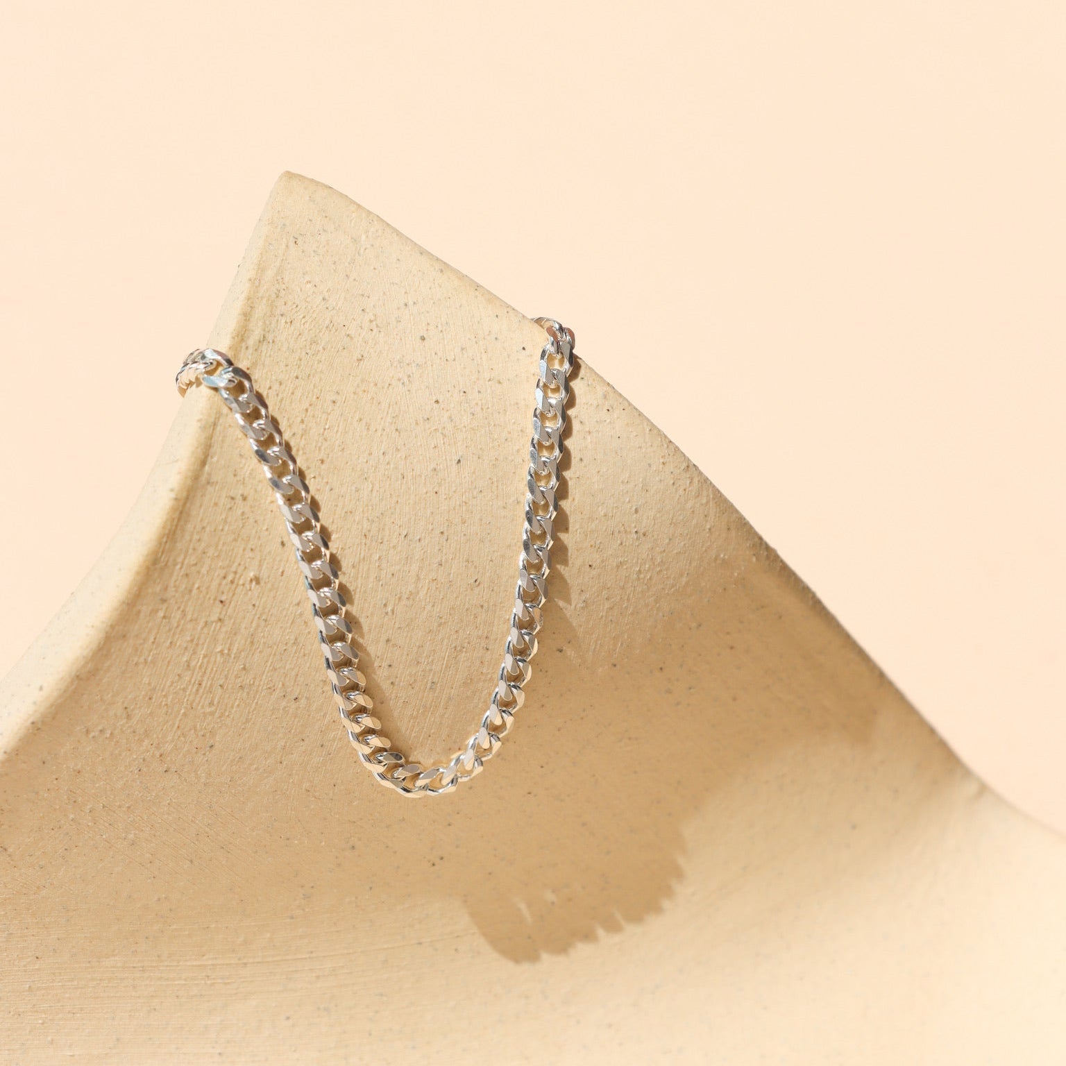 Sterling silver curb link chain draped over a beige textured ceramic prop, set against a warm peach-toned background with gentle sunlight and shadows.