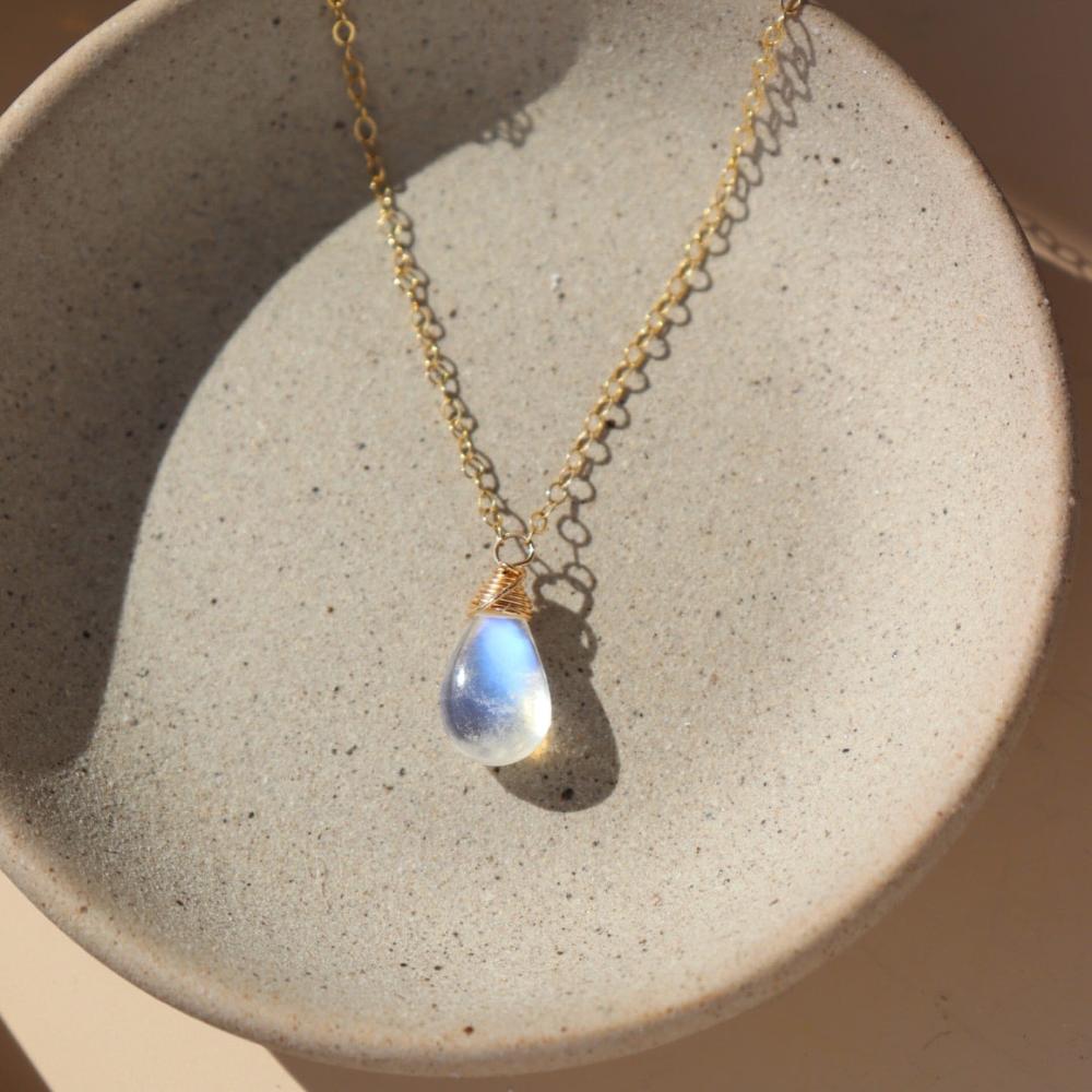 14k gold fill necklace with natural teardrop moonstone full of blue flash laid on a gray moonstone plate.