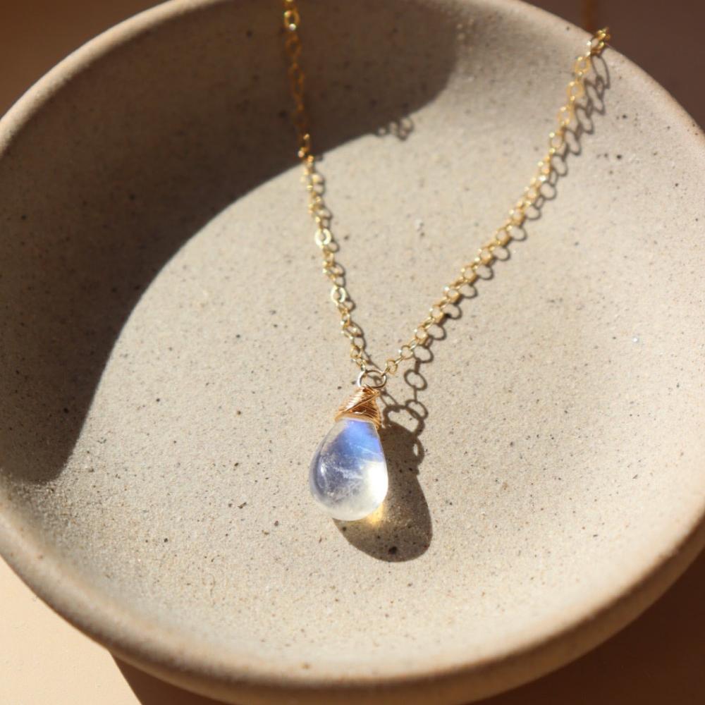 14k gold fill necklace with natural teardrop moonstone full of blue flash laid on a gray moonstone plate.