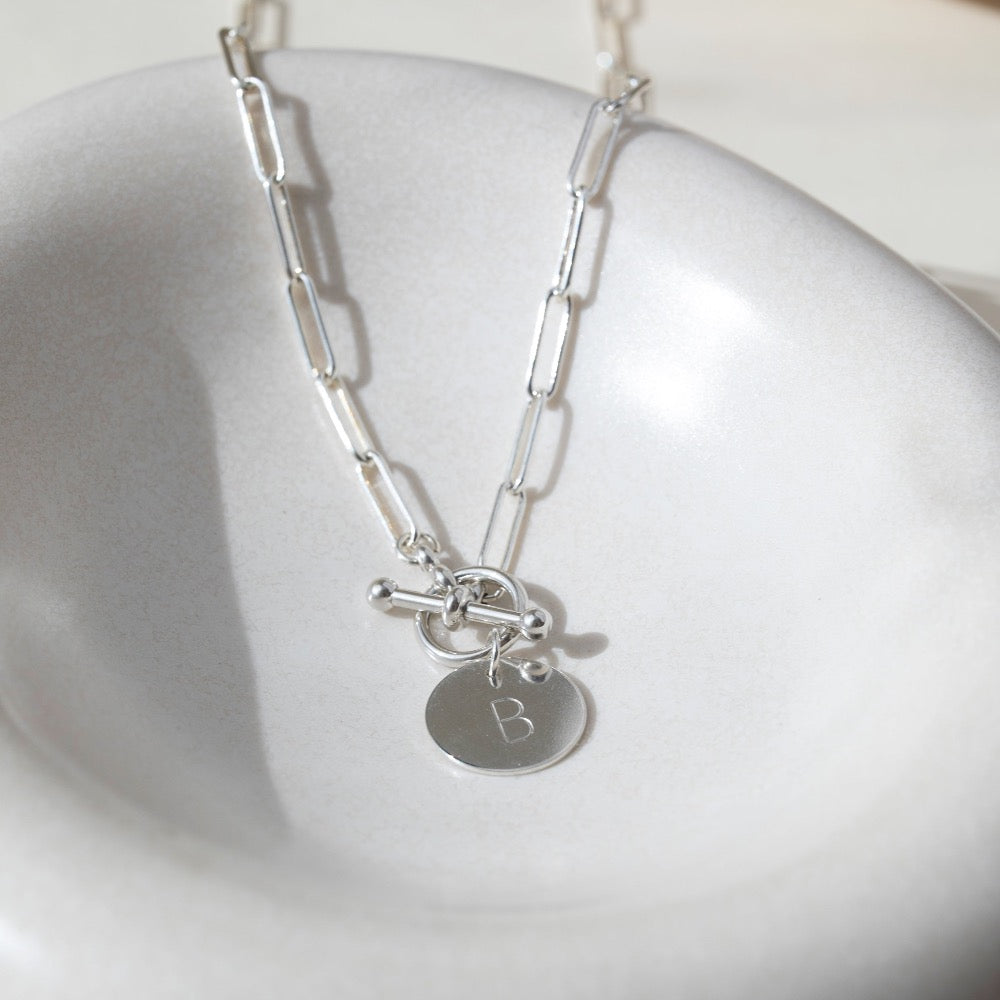Sterling silver chain link necklace with a toggle clasp and monogrammed "B" flat disk charm, photographed on a white dish
