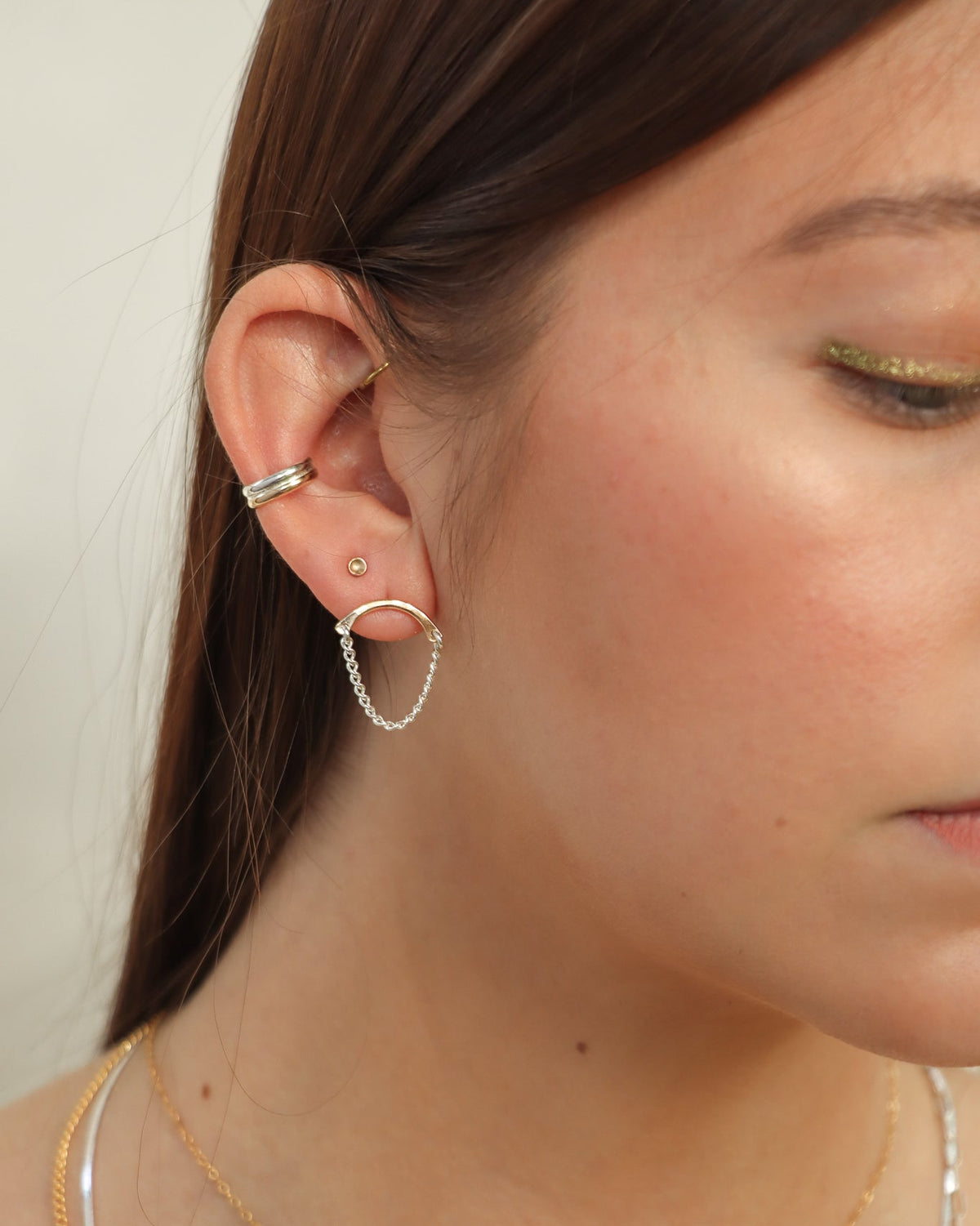 Close-up of a model wearing mixed-metal earrings with a gold curved bar and silver chain drop, styled with layered necklaces and ear cuffs.