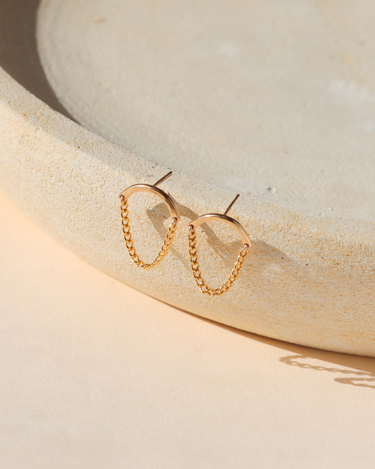 Pair of gold chain drop earrings featuring curved gold bars connected by delicate draped chains, displayed on a textured cream stone surface in warm natural sunlight.