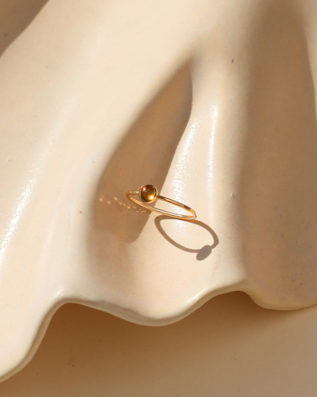 Delicate gold stacking ring with a polished gold ball accent, displayed on a cream ceramic surface in warm natural sunlight.
