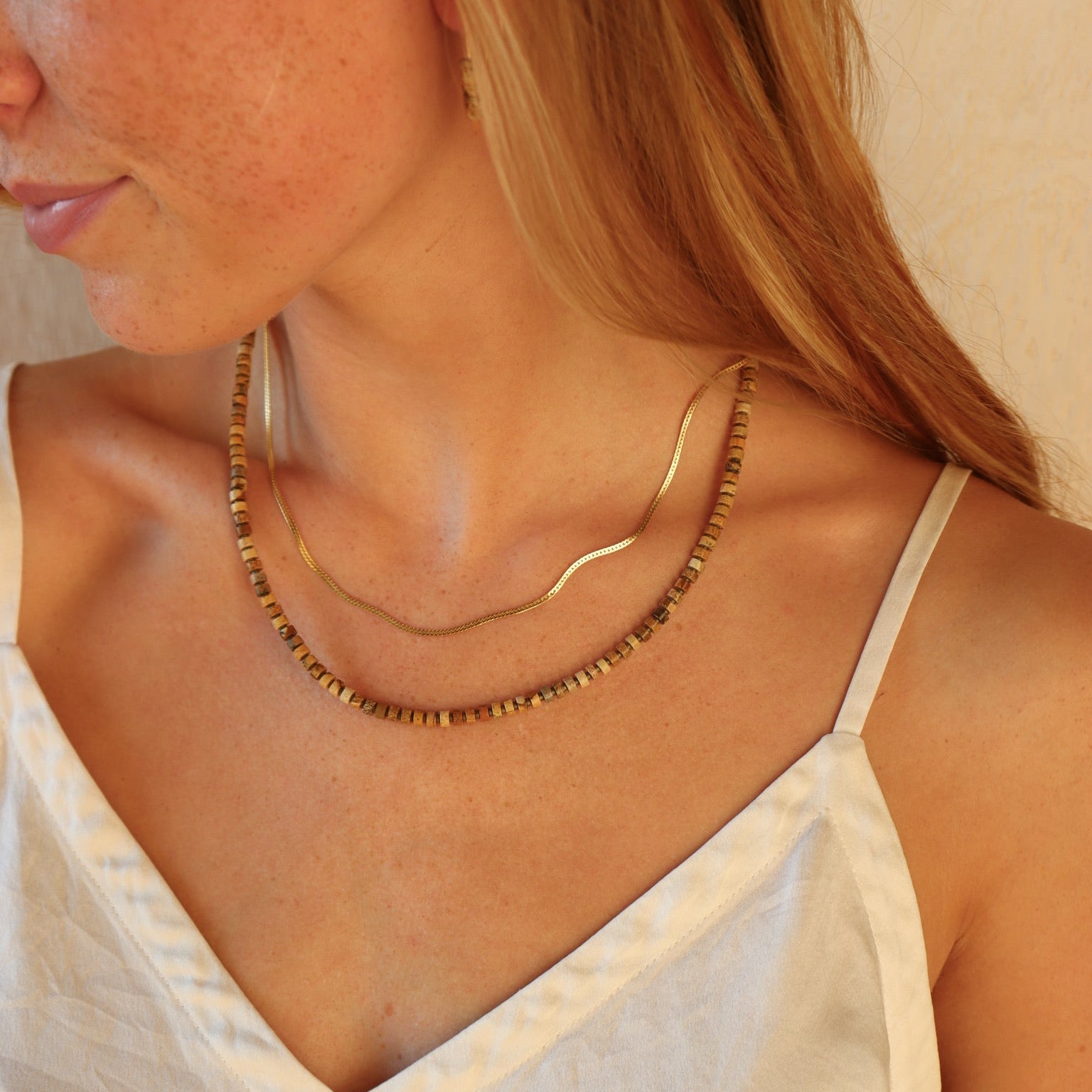 Side profile view of a blonde woman with freckles wearing a white camisole top, showcasing two layered necklaces: a delicate flat gold snake chain layered with a longer strand of Earth-toned square and cylindrical beads.