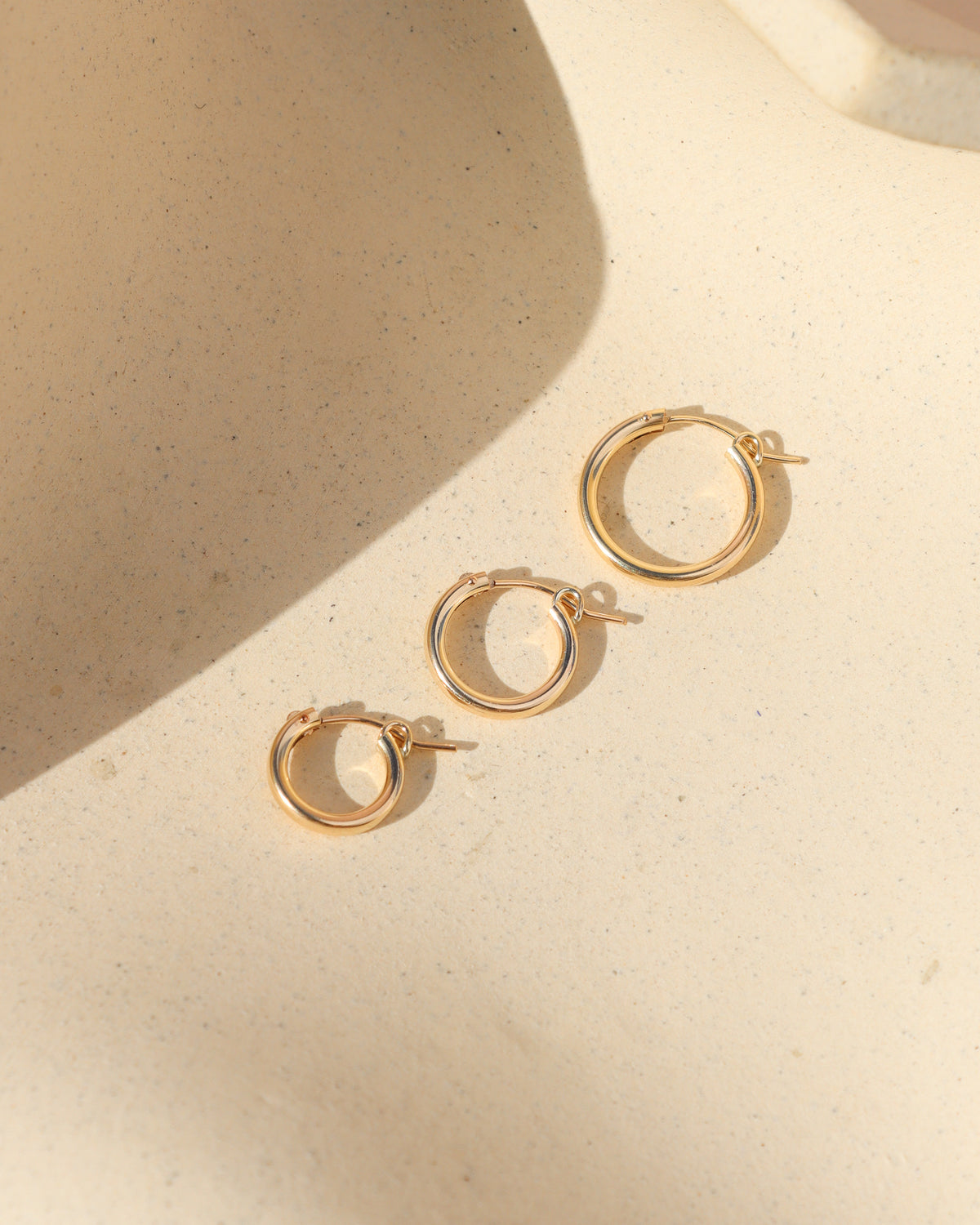 Three polished gold hoop earrings in graduated mini, small, and medium sizes, arranged diagonally on a textured cream surface with warm sunlight and soft shadows.