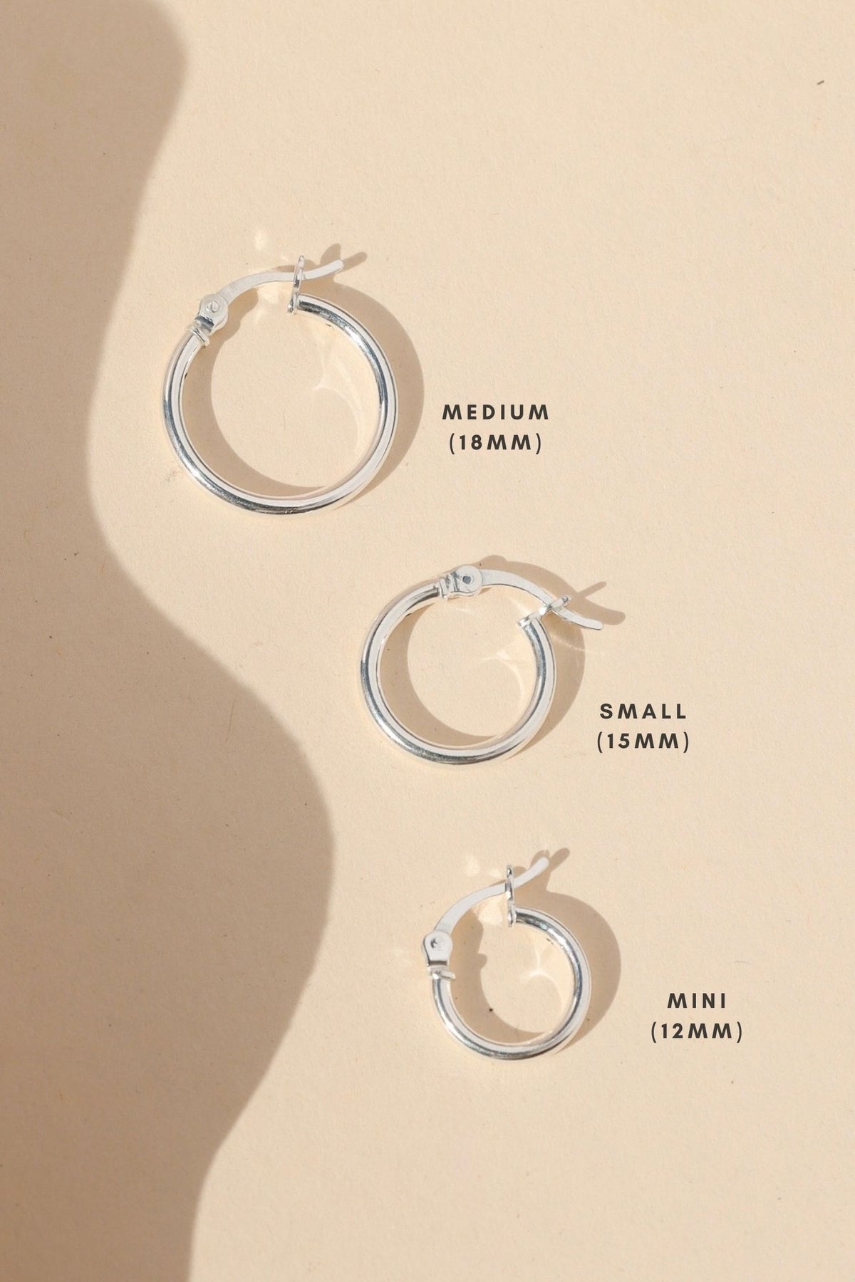 Three polished silver hoop earrings in medium (18mm), small (15mm), and mini (12mm) sizes, arranged on a warm cream background with soft sunlight and shadows to show size differences.
