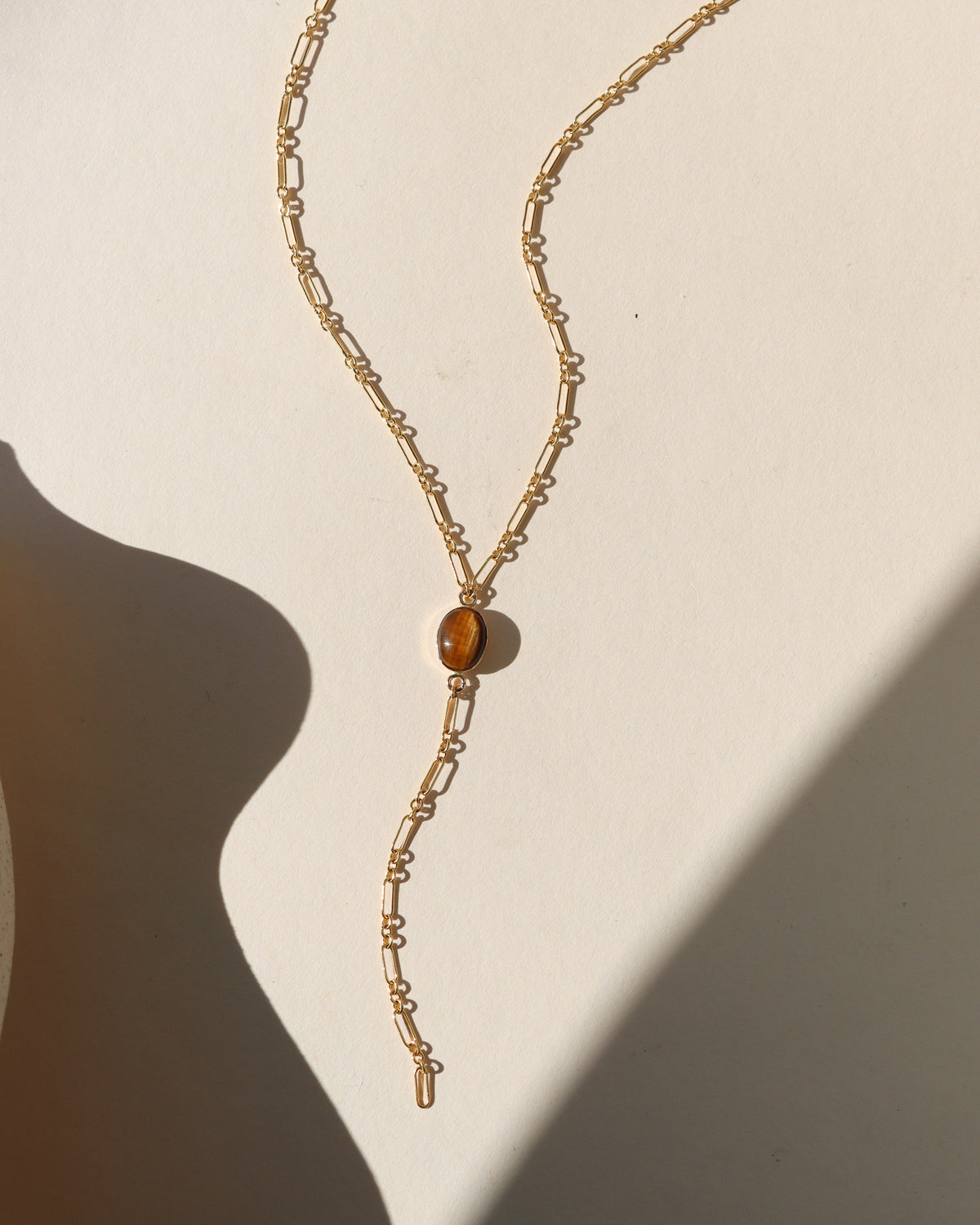 Gold Y-necklace featuring an oval tiger’s eye gemstone and elongated paperclip chain drop, displayed in warm sunlight.