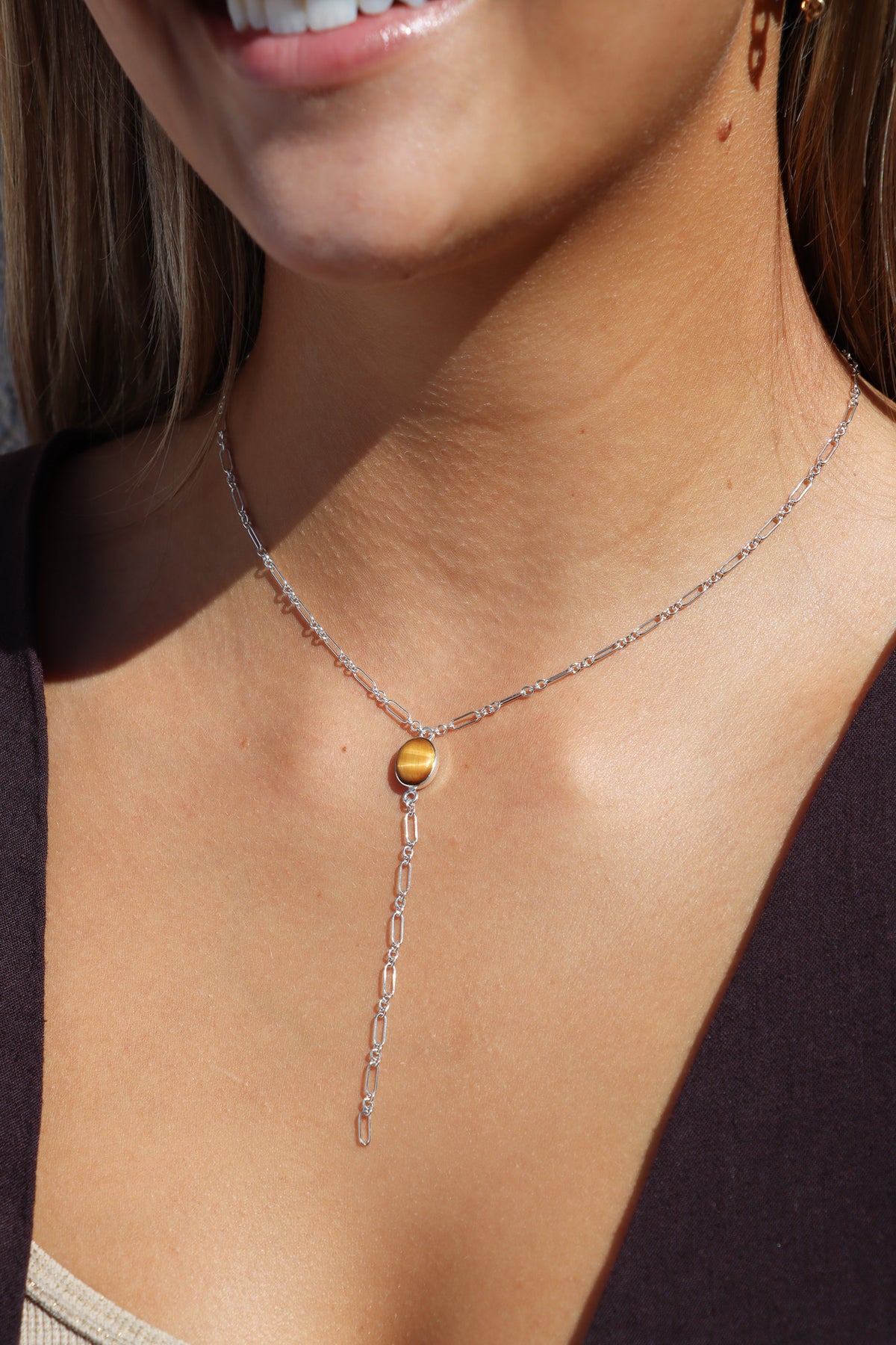 Silver Y-necklace with an oval tiger’s eye gemstone centerpiece and delicate paperclip chain drop, worn in warm sunlight.