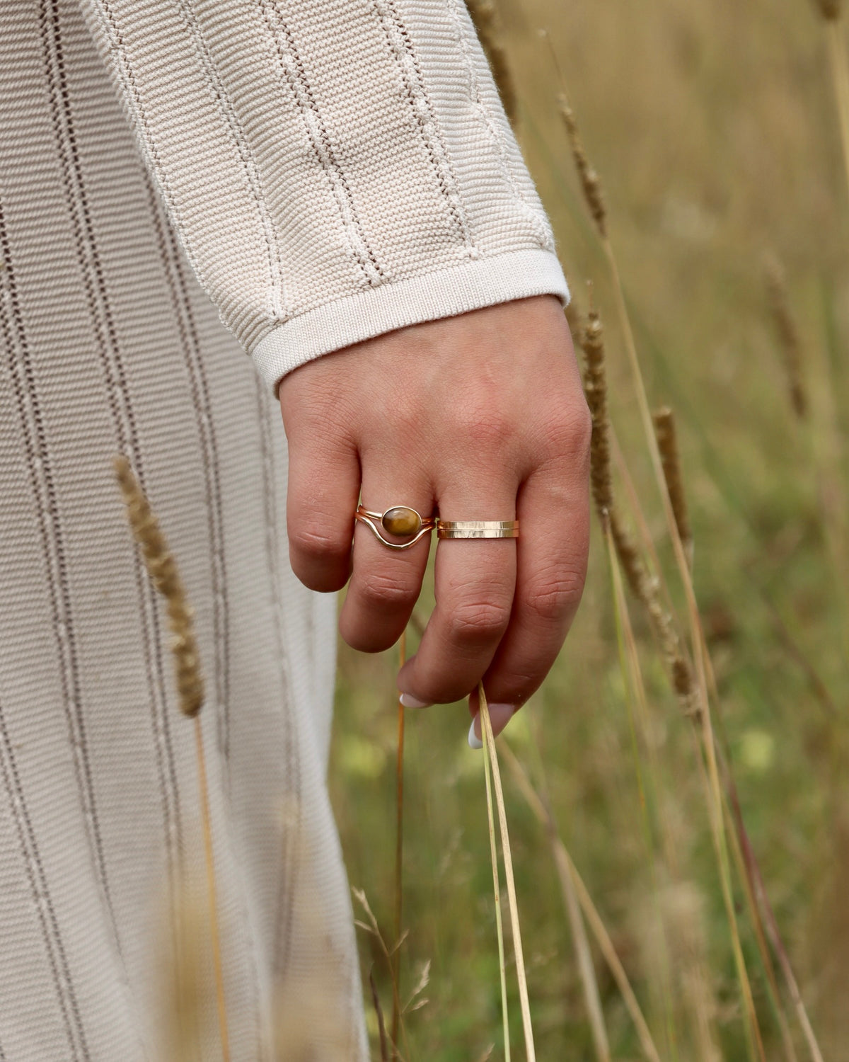 Hand wearing a tiger’s eye gemstone ring and a textured gold band, styled with a cream knit dress outdoors.
