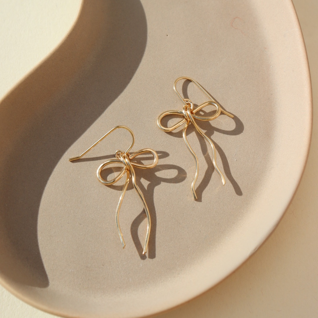 delicate 14k gold fill bow earring hand made by Token Jewelry, photographed on a ceramic dish in the sunlight