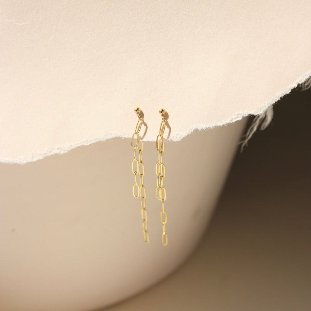 A 14k gold fill small paper clip chain, dropped from a small gold ball stud, the earrings are displayed on a cream backdrop in the sunlight 