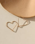 14k gold fill open heart slide earrings. This earring features a gold wire that is shaped into an open heart, with a hook closure. Earrings are displayed on a cream backdrop, rested on a tan dish. 