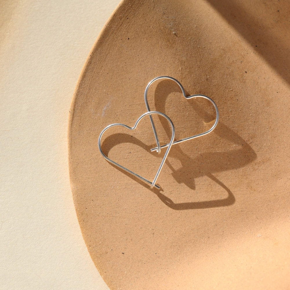 sterling silver open heart slide earrings. This earring features a silver wire that is shaped into an open heart, with a hook closure. Earrings are displayed on a cream backdrop, rested on a tan dish. 