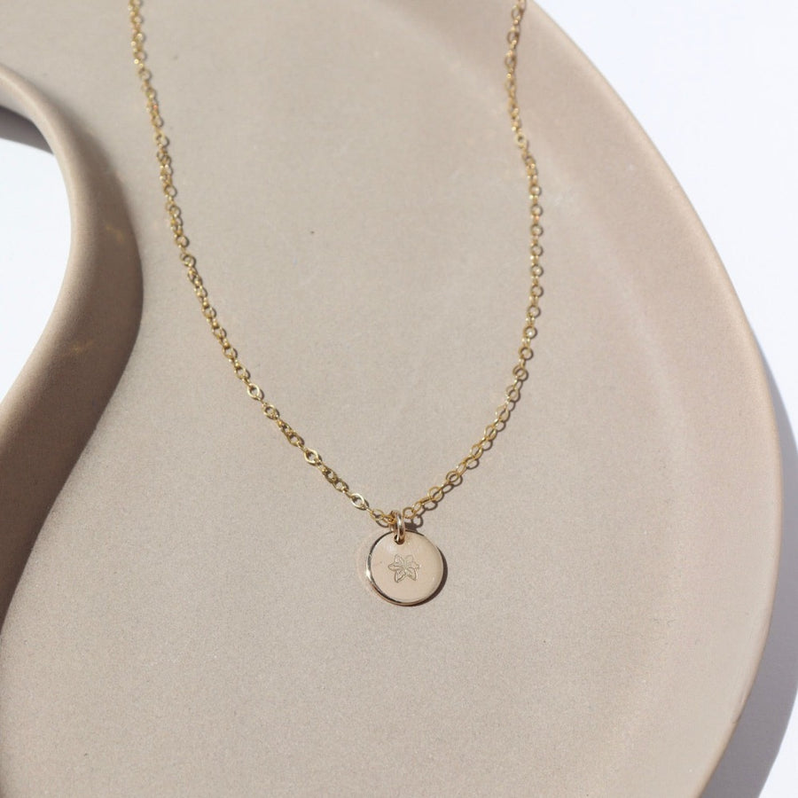 14k gold fill Tiny Birth Flower Disc Necklace laid on a white plate in the sunlight. This necklace features our simple chain with a birth flower pendent of your choice.