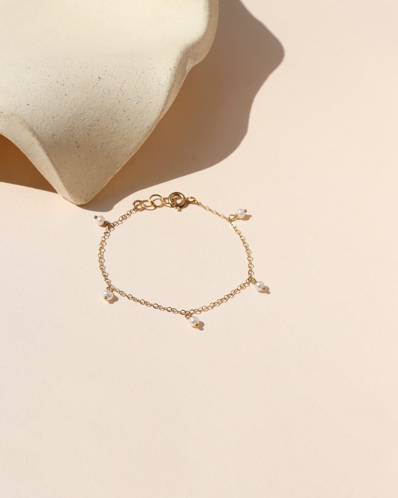 A delicate gold chain bracelet featuring small, dangling white pearls, laid flat on a light beige background alongside a ceramic display piece.