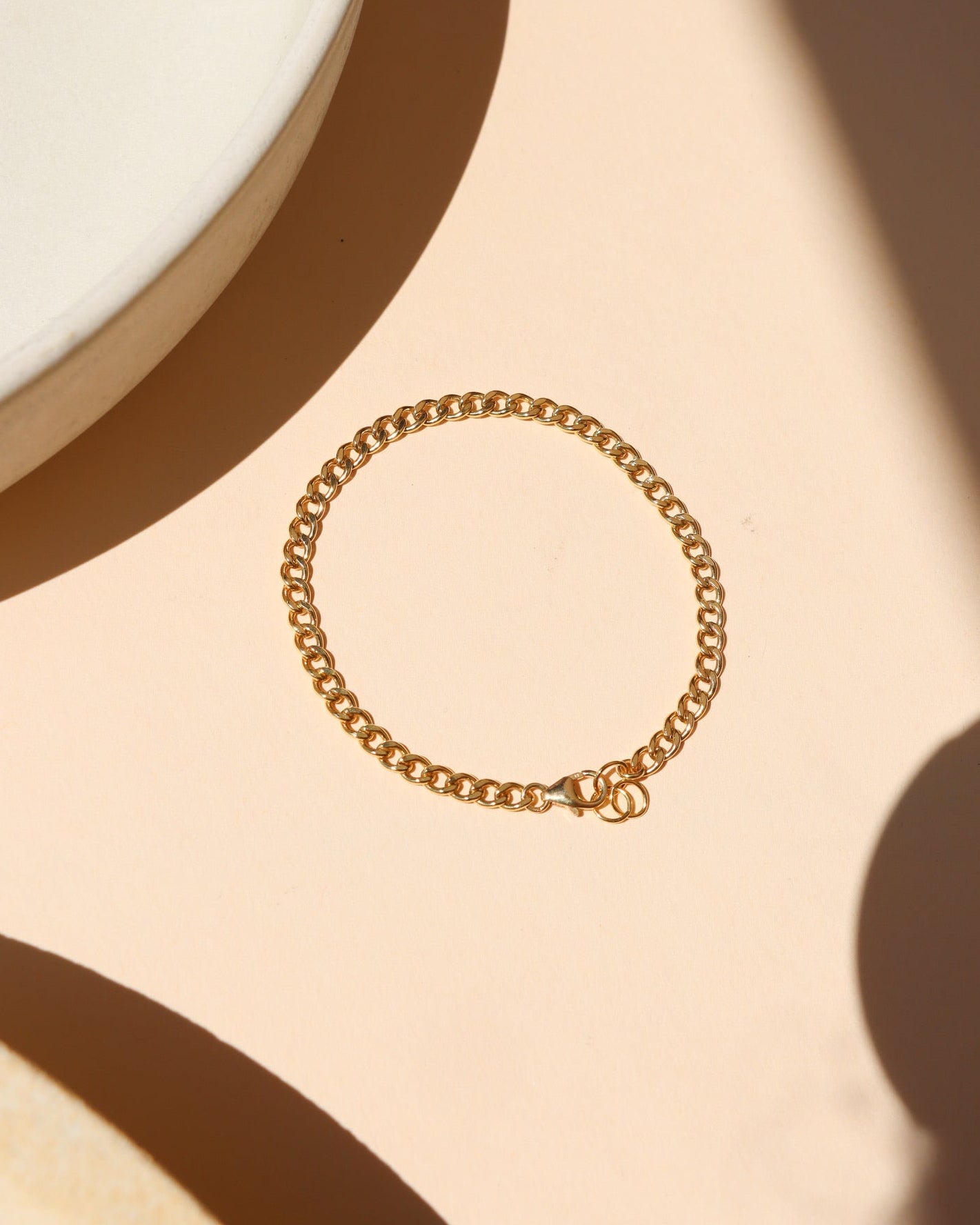Classic gold curb link chain bracelet featuring interlocking flattened links and a spring ring clasp, laid flat on a peach-beige surface near a ceramic dish with warm natural light shadows.