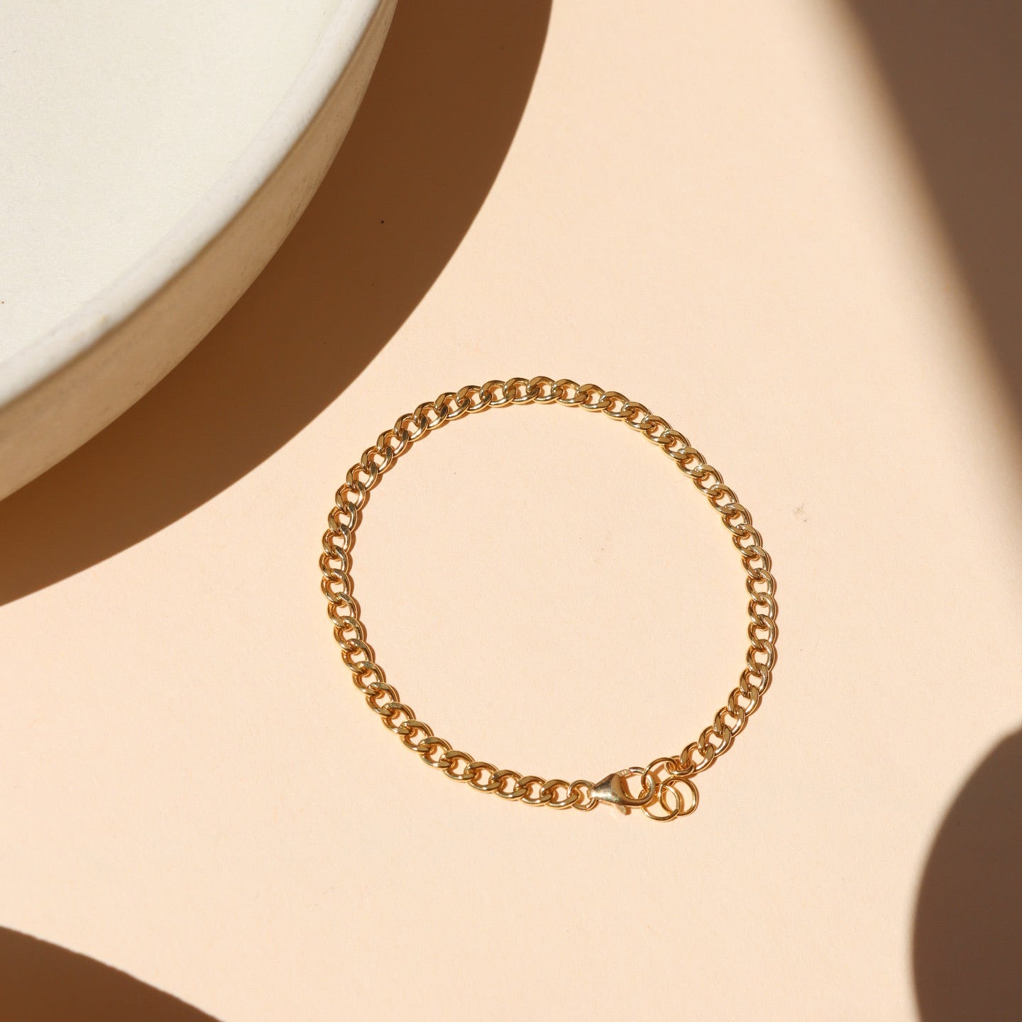 Classic gold curb link chain bracelet featuring interlocking flattened links and a spring ring clasp, laid flat on a peach-beige surface near a ceramic dish with warm natural light shadows.