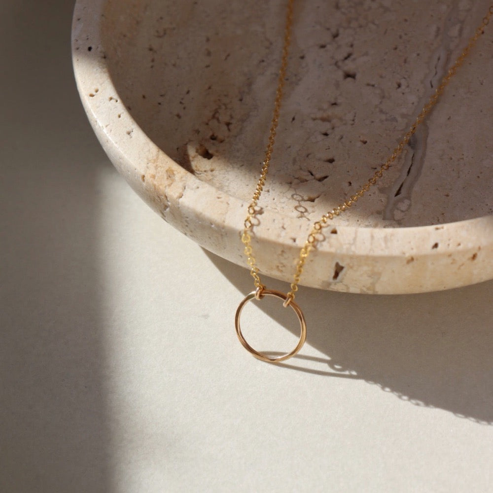 14k gold fill Eternity Necklace placed on a cream colored dish. This necklace and plate is sitting in the sunlight. This necklace features an open circle disc that is attached to our simple chain. Token Jewelry is hypoallergenic and nickel free. - Token Jewelry