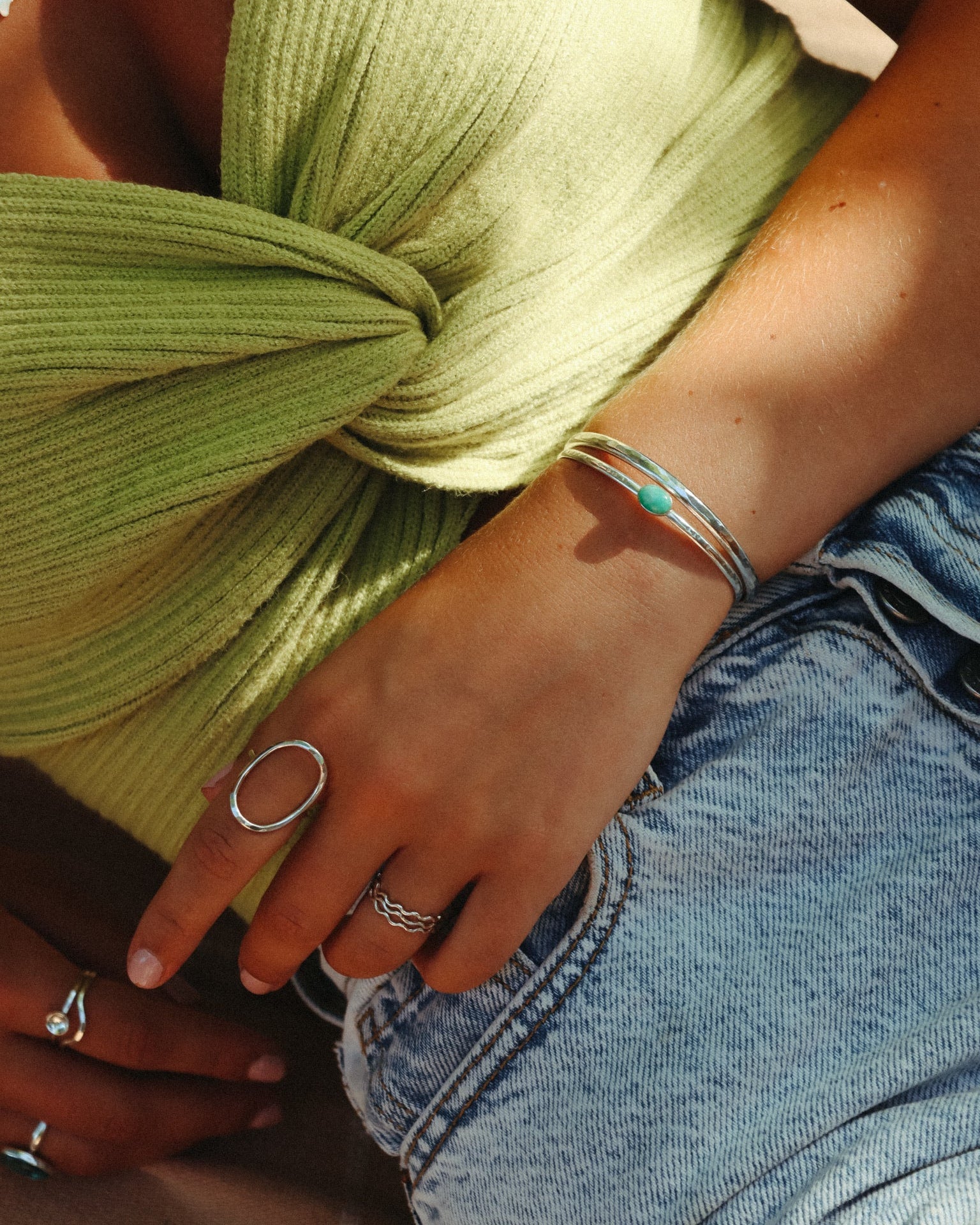 A close-up of .925 sterling silver stacked cuff bracelets—one featuring a small oval turquoise stone—paired with silver statement rings and a green top.