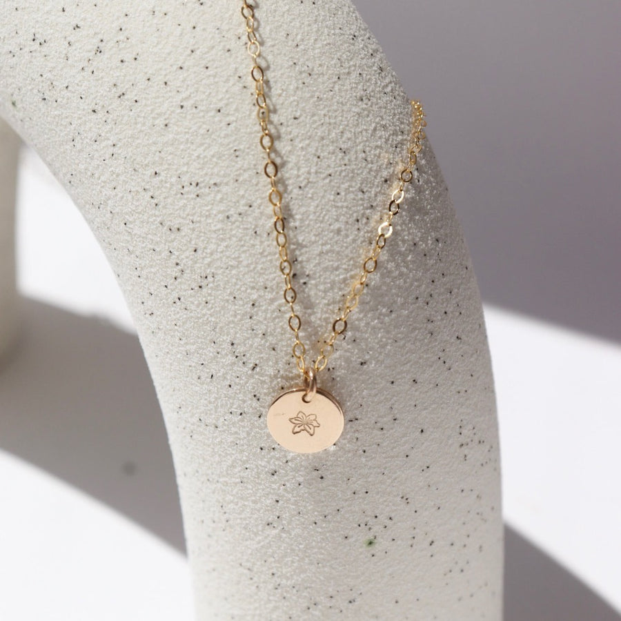 14k Gold fill Children's tiny birth flower charm necklace laid on a white jewelry stand in the sunlight.