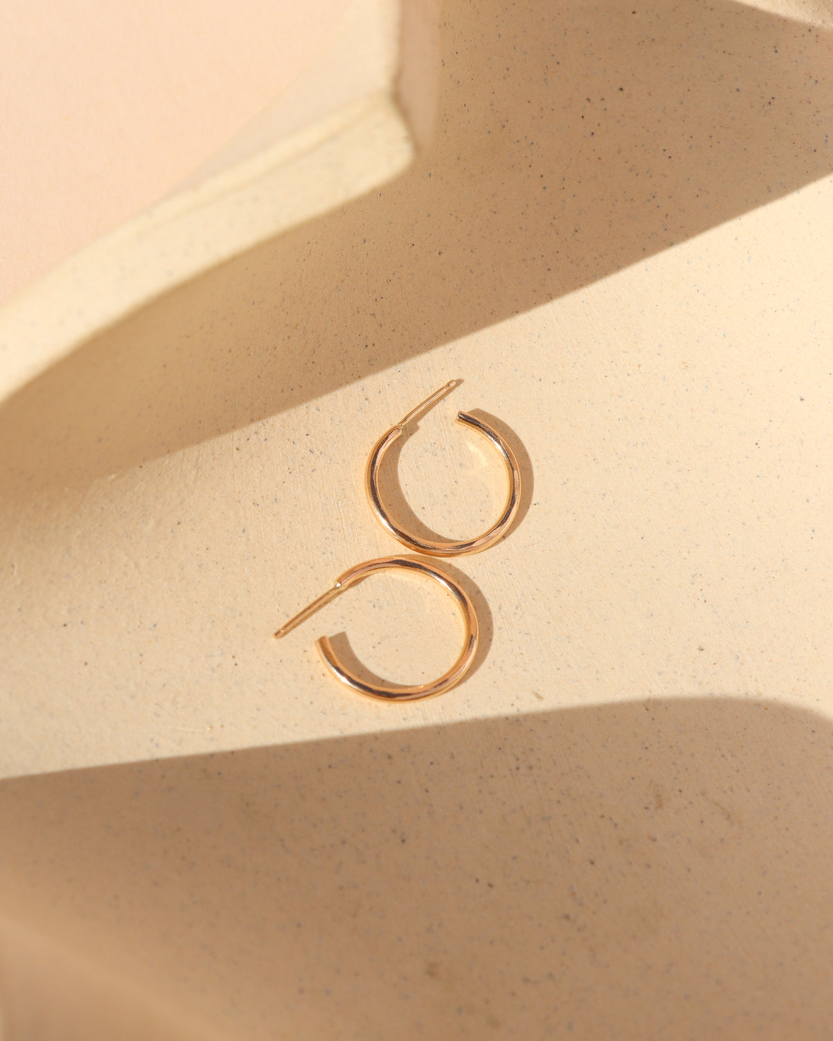 Pair of delicate gold hoop earrings resting on a warm, textured cream surface with soft sunlight and natural shadows.
