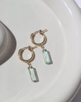 14k gold fill Flynn Hoops laid on a tan plate in the sunlight. These earrings feature a hoop like earring with a dangle of a Fluorite tube gemstone.