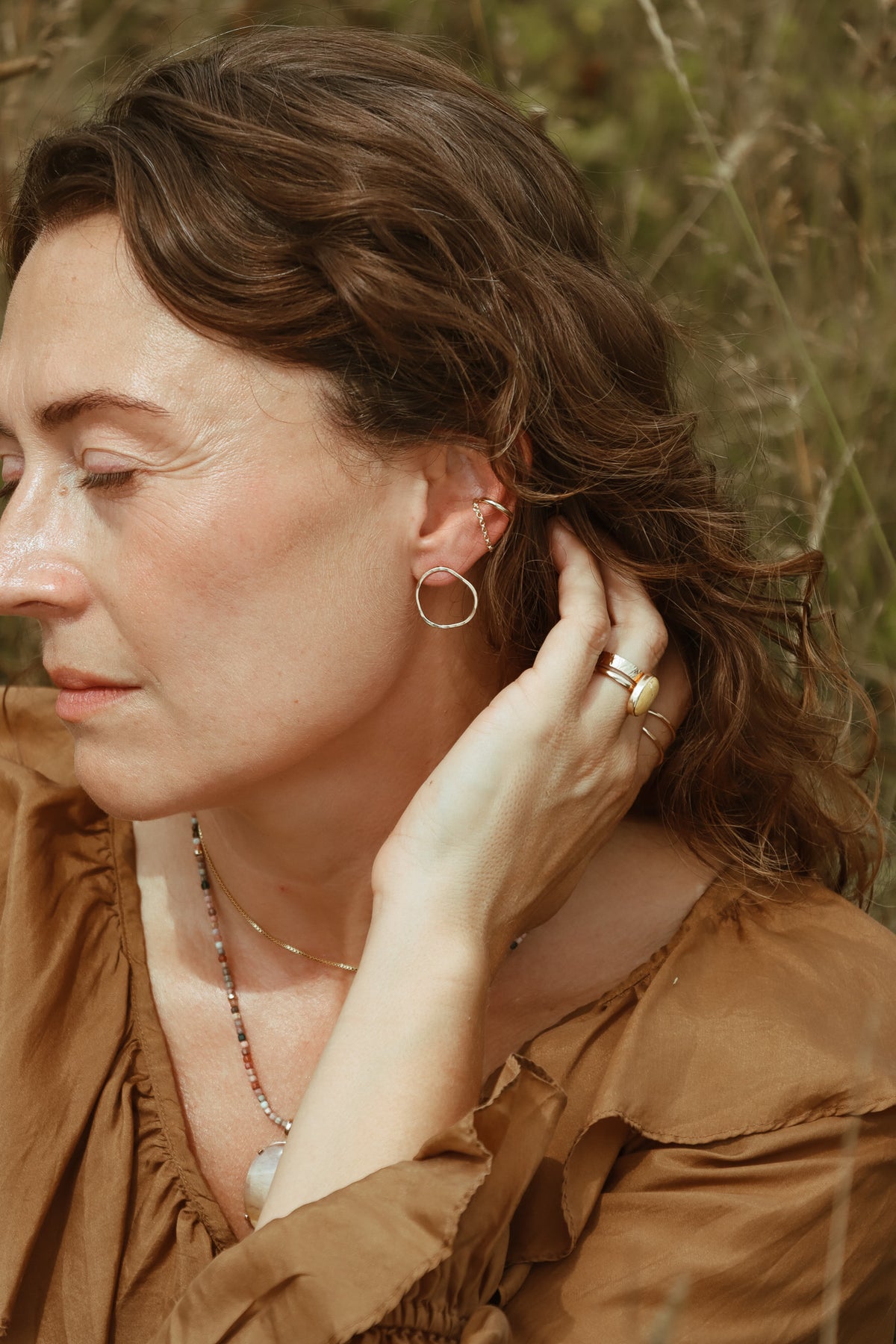 Model wearing a organic shaped stud earring with layered ear jewelry, rings, and necklaces, styled with a brown ruffled blouse.