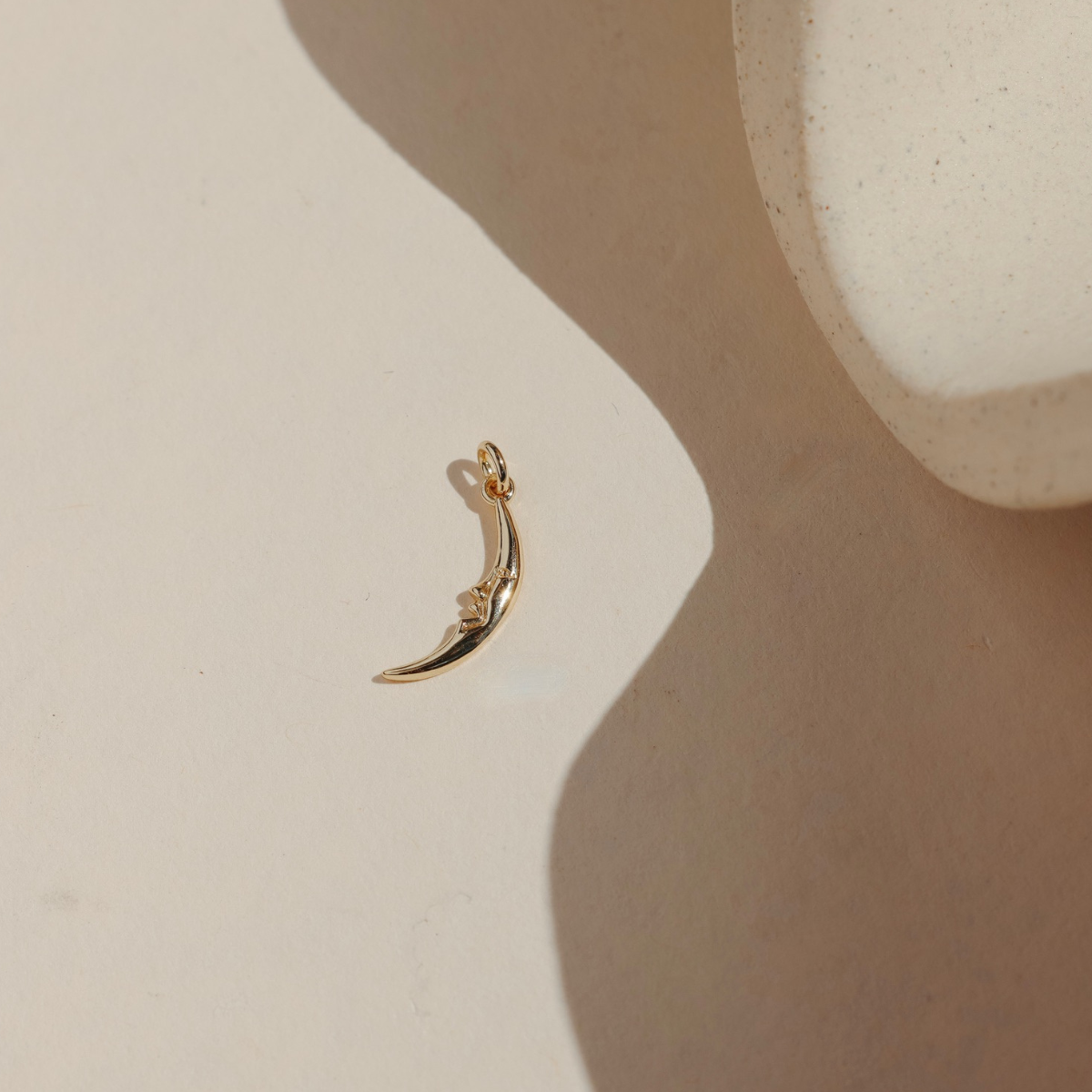 Gold crescent moon charm with a sculpted face, photographed on a neutral stone surface in warm natural light.