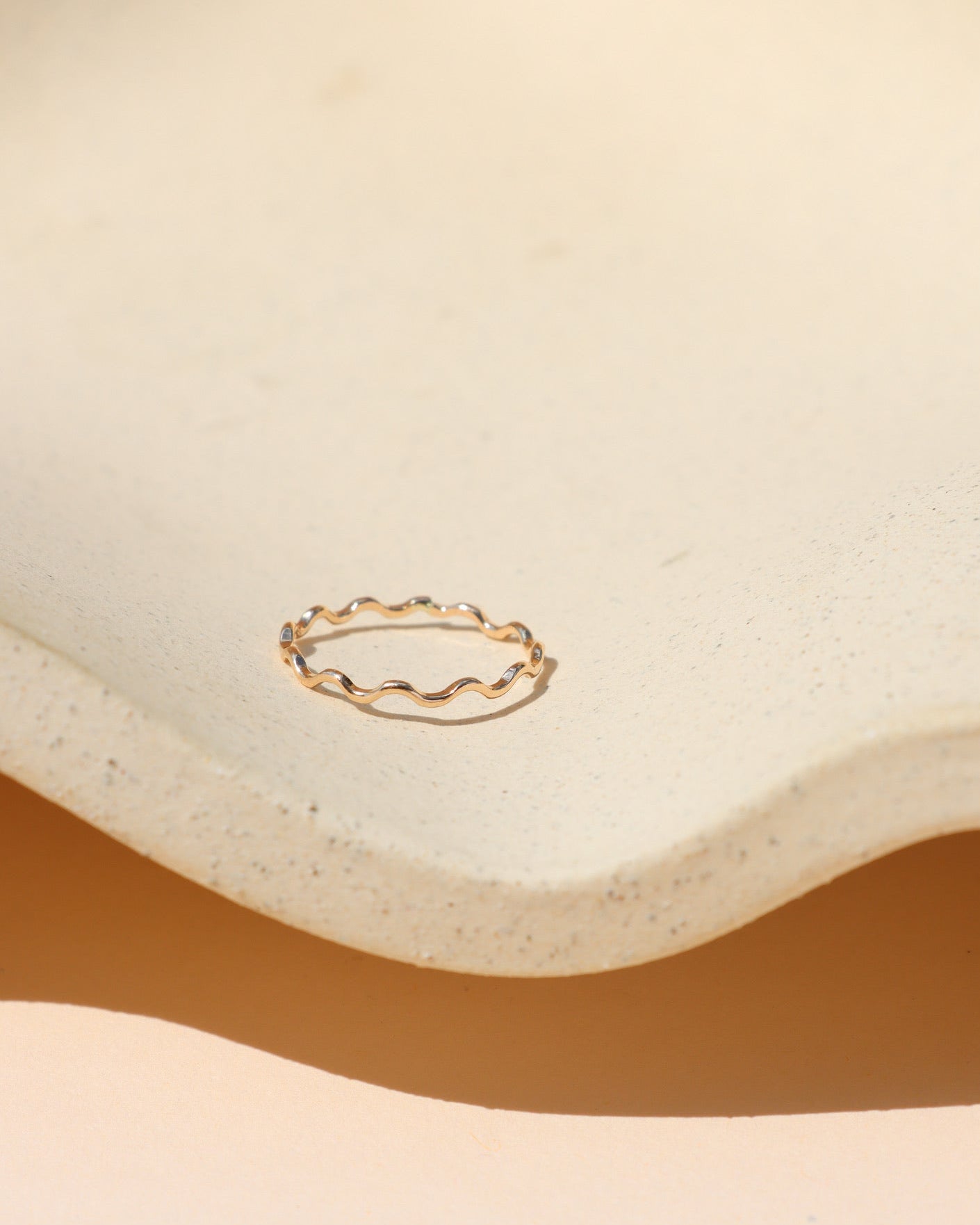 Minimalist gold ring with a gentle squiggly wave pattern, displayed inside a cream-colored textured ceramic dish with soft natural sunlight and warm shadows.